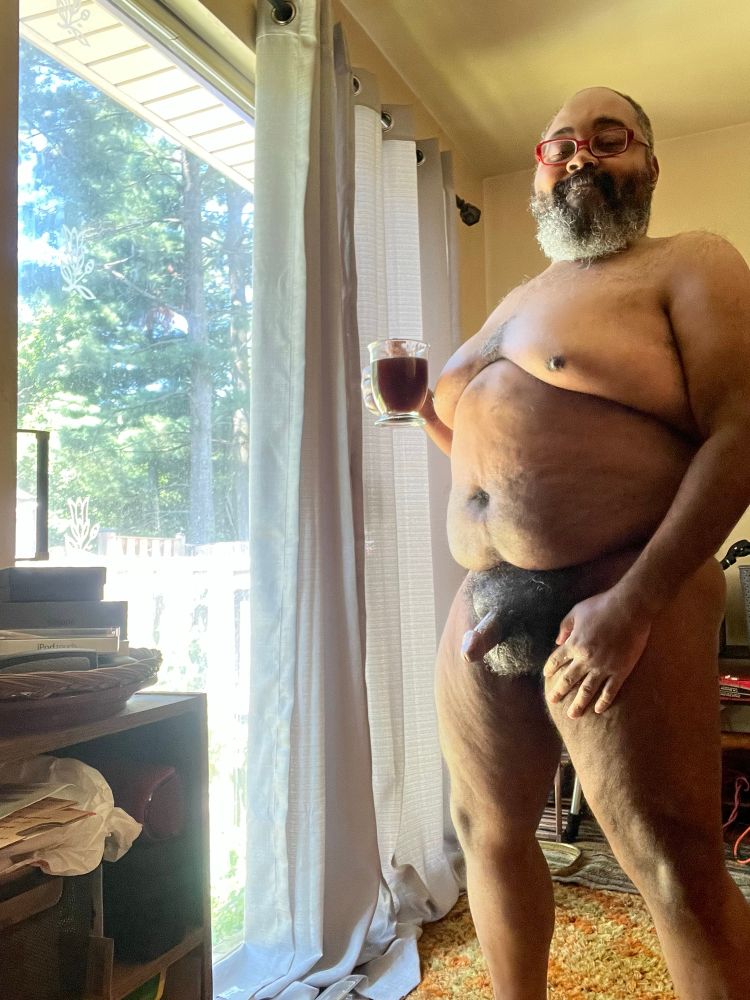 Bearded Black man: he stands in his living room by a sliding glass door on a sunny day. He is fully nude, and in his right hand is a clear mug of black coffee. His other hand is resting on his thigh. He is turned 3/4 to the camera and is smiling. He is fluffed.