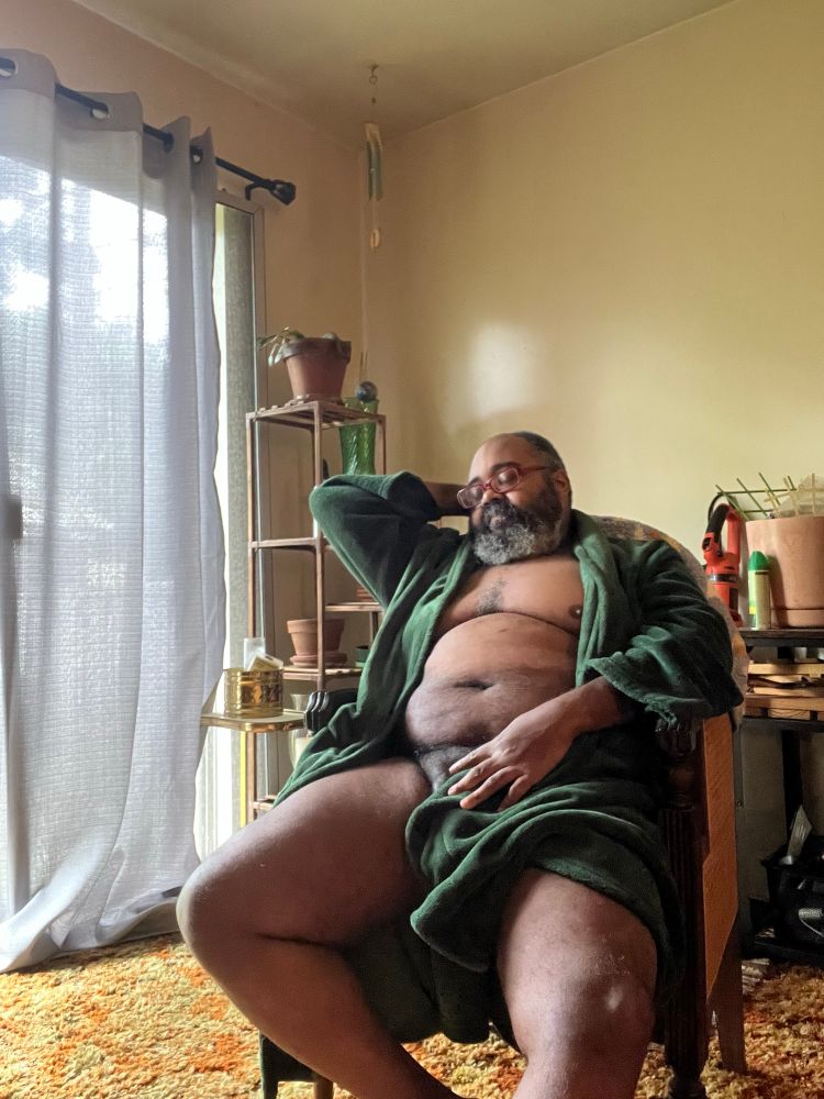 Bearded Black man: he’s sitting in a wooden chair with wicker panels and in his living room. He is wearing a green plushy robe that is open to reveal his chest and thighs. His one hand obscures his genitals while the other is behind his head. He is otherwise nude. There is faint light from the sliding glass window covered by a blue sheer curtain. His face is serious and with his eyes closed.
