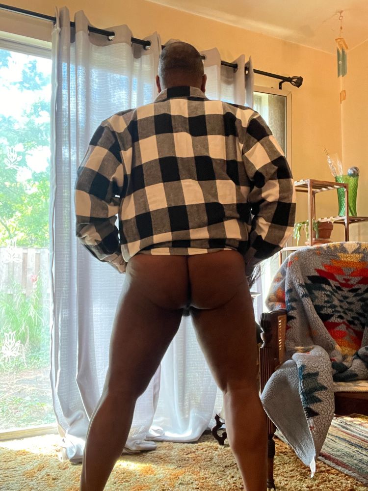 Black man wears a Buffalo plaid shirt in white and black. He stands in his living room with back facing the camera and shirt hiked up to his waist. He is not wearing any bottom clothing and his bare bum is visible.