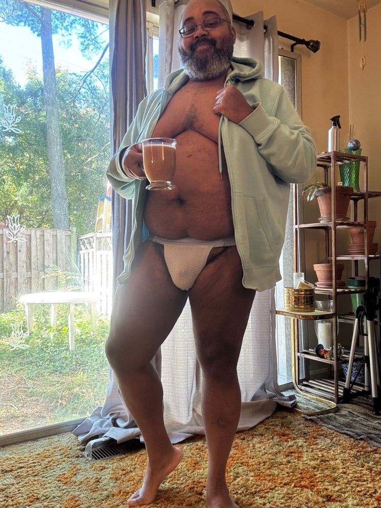 Bearded Black man: he stands nearly naked in his living room before a sliding glass door with blue sheer curtains partially open. He wears a powder blue hoodie, open, and a powder blue jockstrap. He has one leg slightly bent; he holds a clear mug of cafe au lait in one hand and the zipper edge at the collar in the other.