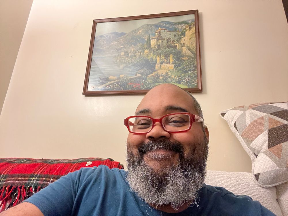 Bearded Black man: he’s in a teal blue tee and is sitting in a living room with a mass print of the Italian coast and a castello by the sea.