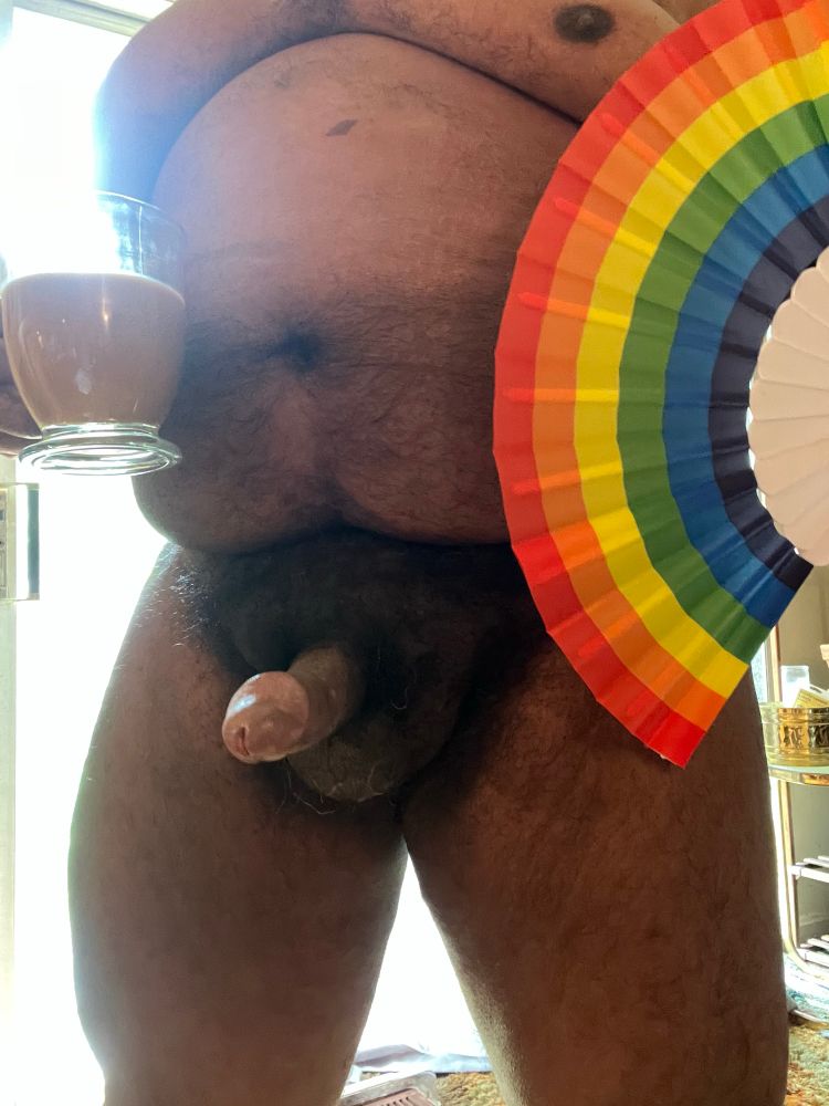 Black man: his nude body visible as sunlight from a sheer curtain glow behind him. In one hand is cafe au lait in a clear glass mug, and in the other is a rainbow hand fan with white ribs. His penis is erect. His face is not visible.