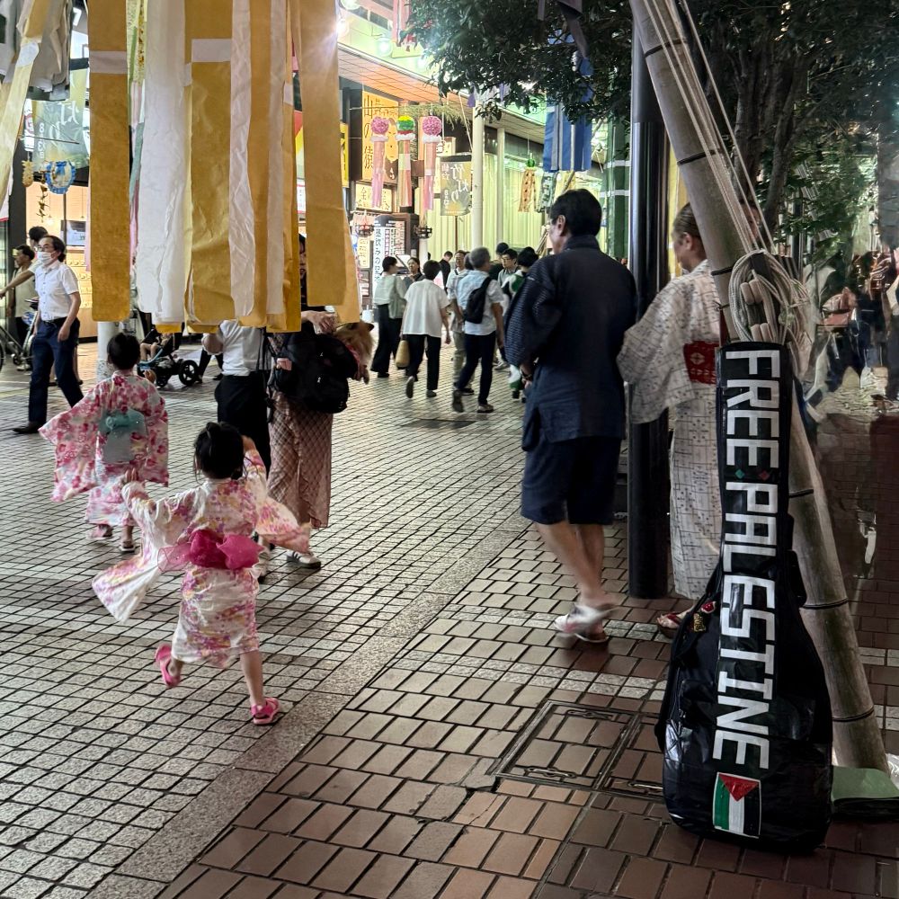 A placard saying Free Palestine stands in a corner of downtown. It is Tanabata Festival Day, so some people wearing Yukata (a kind of Japanese Kimono) were walking in the street, including children running and playing.