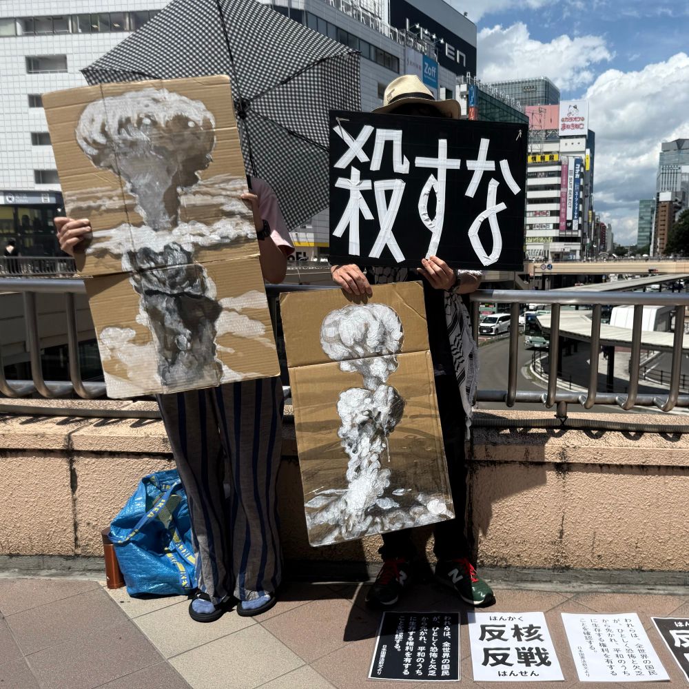 路上に立つ人物ふたり。ひとりは、長崎に投下された原子爆弾「ファットマン」のきのこ雲が描かれたプラカードを持っている。もうひとりは、広島に投下された原子爆弾「リトルボーイ」のきのこ雲が描かれたプラカードを右手に、左手には「殺すな」と大書されたプラカードを持っている。その足元には、「反核反戦」や「われらは、全世界の国民が、ひとしく恐怖と欠乏から免れ、平和のうちに生存する権利を有することを確認する」と書かれたプラカードも置かれている。青く晴れた空の下。