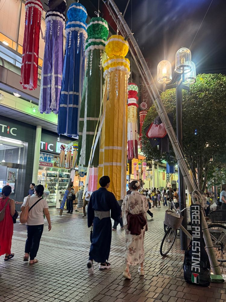 A placard saying Free Palestine stands in a corner of downtown. It is Tanabata Festival Day, so some people wearing Yukata (a kind of Japanese Kimono) are walking in the street, including young lovers holding their hands each other.
