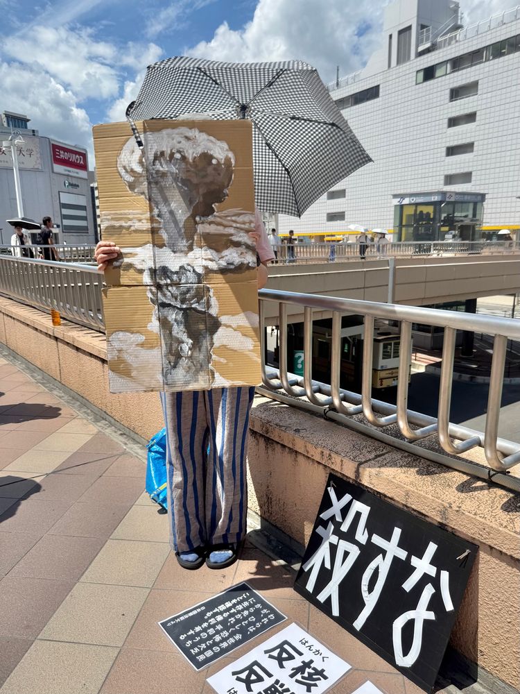 路上に立つ人物。長崎に投下された原子爆弾「ファットマン」のきのこ雲が描かれたプラカードを持っている。足元には、「殺すな」「反核反戦」「われらは、全世界の国民が、ひとしく恐怖と欠乏から免れ、平和のうちに生存する権利を有することを確認する」と書かれたプラカードも置かれている。青く晴れた空の下。