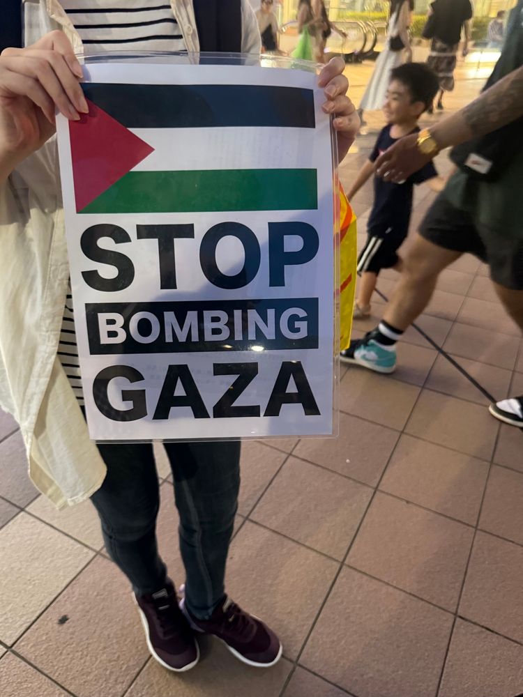 "Stop Bombing Gaza" と書かれたプラカードを持って、街角に立つひと。