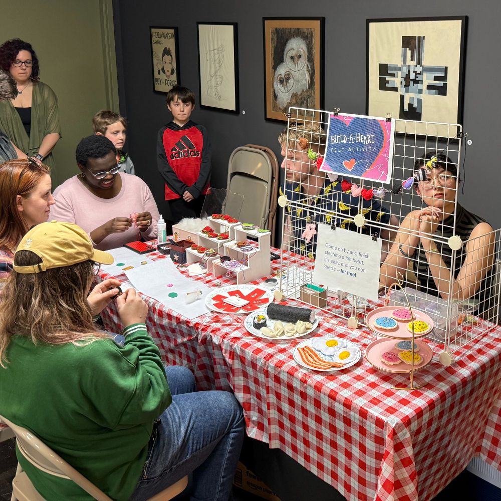 Children and adults stand and sit around a rectangular table dressed with a red and white checkerboard tablecloth with felted objects such as bacon and eggs, cookies, and strawberries on plates sitting on top. Two artists sit behind the table leading a felting activity. 
