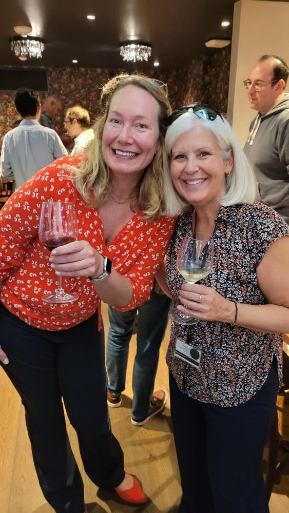 Kim Franklin and Laura Henneman pose together, holding wine glasses. Mark Dredze crosses in the background.
