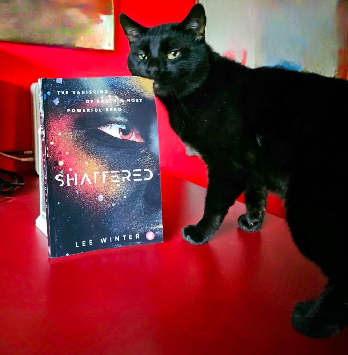 black cat on red table with a paperback version of Lee Winter's "Shattered" looking very annoyed because he is aware that his weekend plans are just being shattered, because I'm going to read it in one sitting.