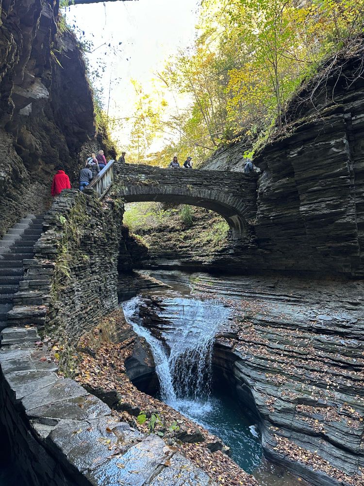 Stone steps on the left leading up to a stone bridge over a carved gorge. The water cascades into a small waterfall & the rock wall is striated.