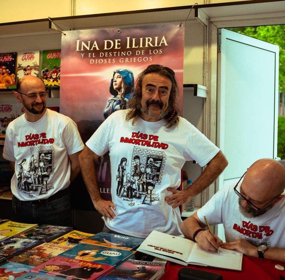 De izquierda a derecha: Javier Ortiz, de la editorial Drakul, junto al guionista Eliseo García y el dibujante Javier Urrea, que firma un ejemplar de "Días de inmortalidad", cómic obra de ambos que ha publicado Drakul. Los tres visten camisetas que reproducen la portada del cómic y están en la caseta de la editorial en una feria del libro.
