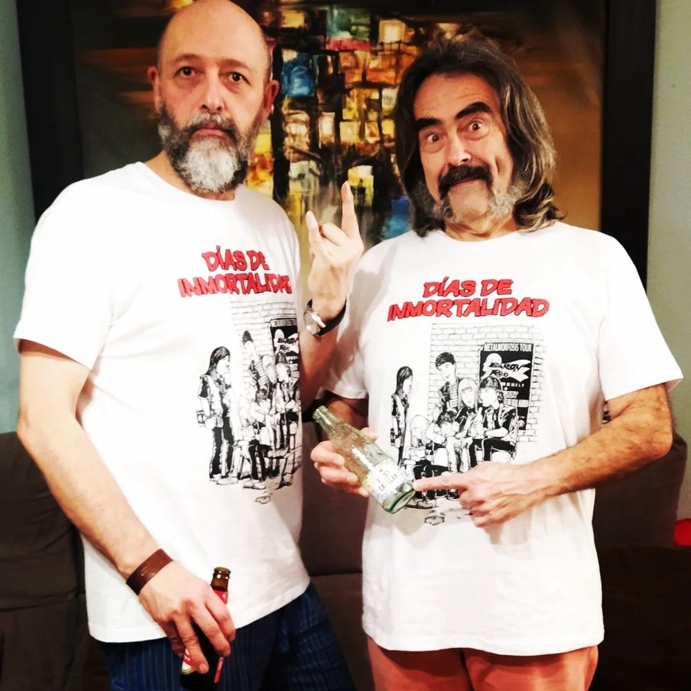 El dibujante Javier Urrea, cincuentón calvo y barbudo, y el guionista Eliseo García, cincuentón patilludo, bigotón y melenudo, autores del cómic "Días de inmortalidad". Posan con camisetas promocionales que reproducen la portada del cómic. La portada reproducida en las camisetas blancas incluye el título "Días de inmortalidad" en letras rojas y una ilustración algunos de los protagonistas del cómic. Son media docena de chicos adolescentes heavies de un barrio madrileño de los años 80, sentados en el banco de un parque.