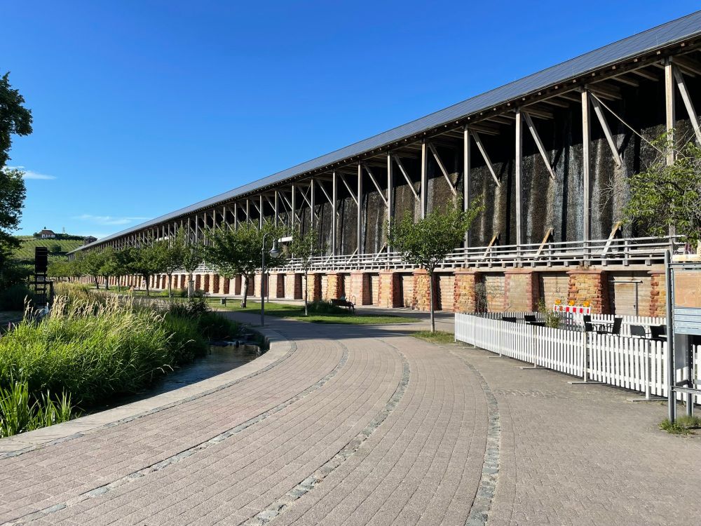 Angled view of most of one side of the Gradierwerk, showing an extremely long wooden structure with dark wooden panels, at the far end a hill with grapevines and a small white chapel, in the foreground a curving stream with reeds and a curving pathway.