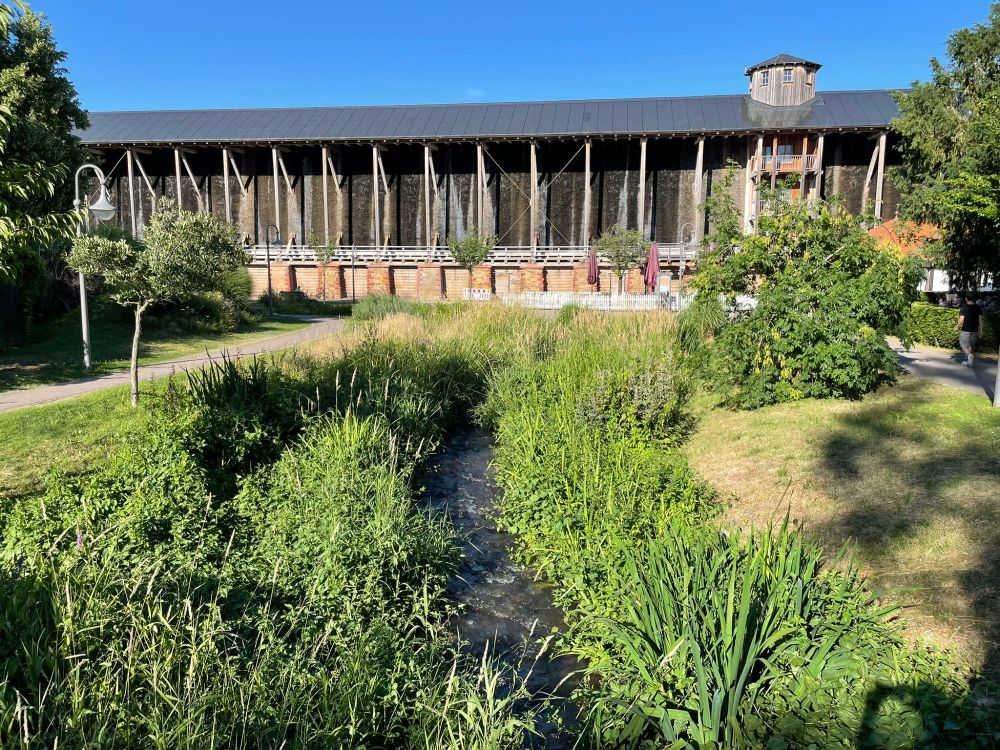 Under a blue sky, a view of one side of the Gradierwerk, a wooden frame with a raised platform and dark panels, her with a small wooden turret, in the foreground a stream with plants either side. 