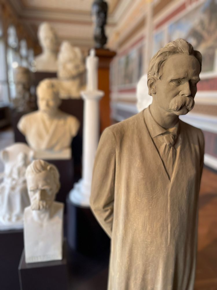 Selection of busts and statuettes of Friedrich Nietzsche in a museum on stepped black pedestals. Close to the viewer, a standing statuette in grey plaster, behind out of focus largely white busts.