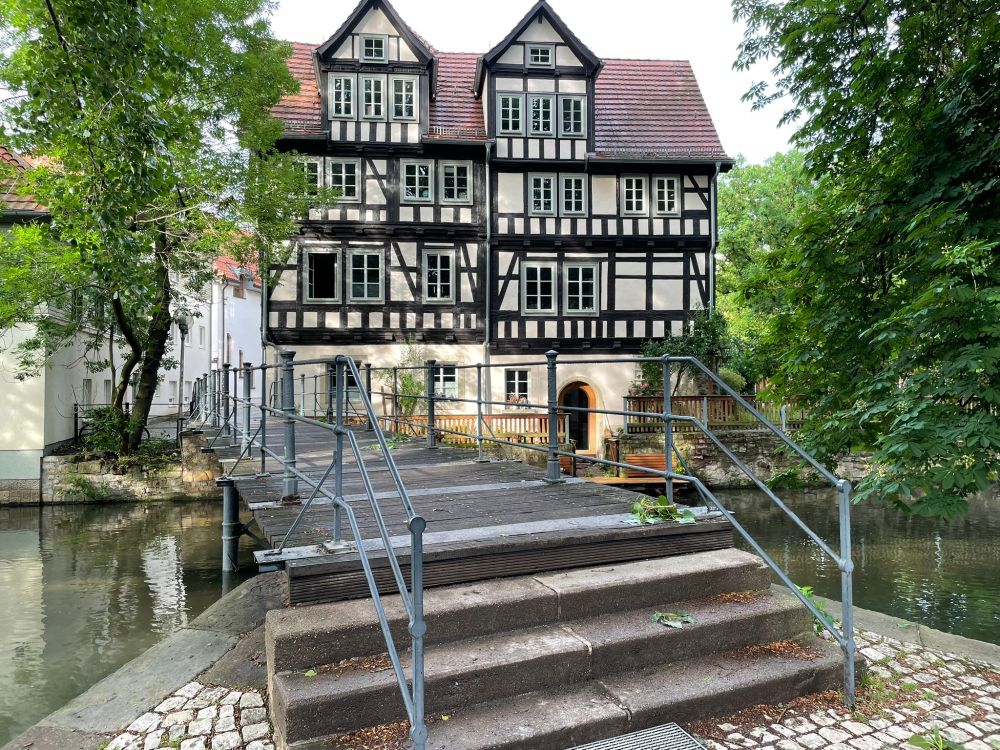 Two half-timbered cream houses with sage window frames with trees either side overlooking a canal with a footbridge of stone steps and wooden boards.
