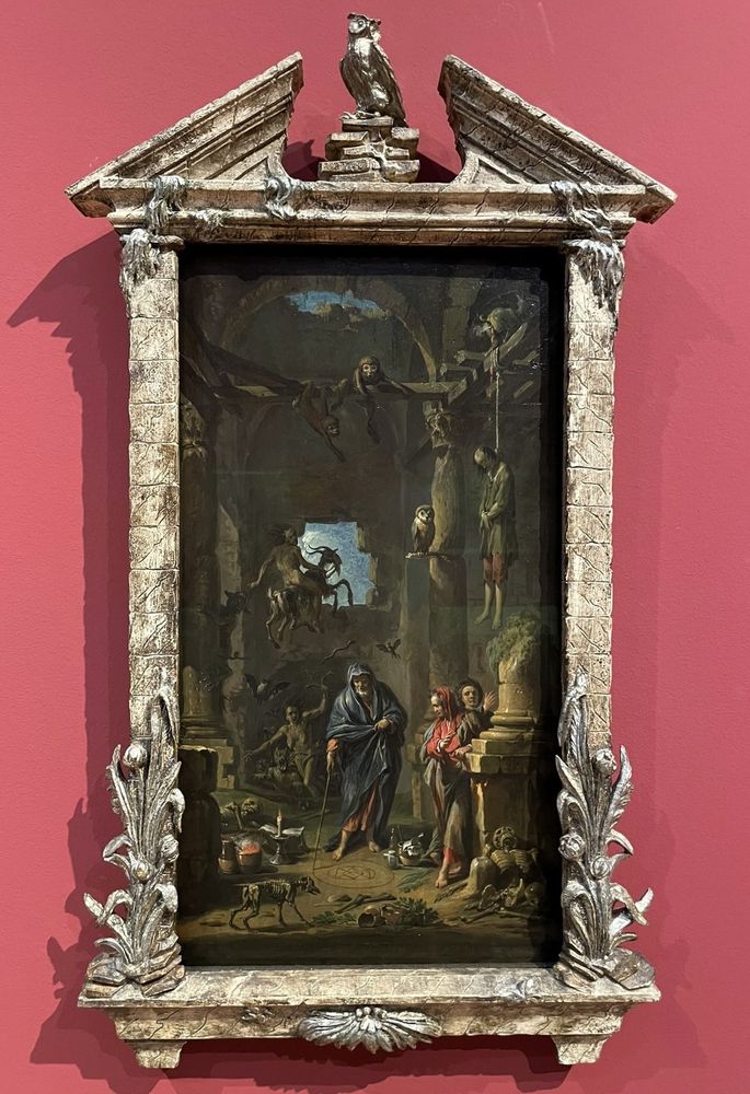 Against a mulberry coloured wall, a carved silver-plated frame in portrait format topped by an owl on a stack of books within a pediment, with plants and flowers at the bottom left and right. The oil painting within the frame shows a sorcerer in a blue cloak reading from a book on the ground, tracing a sigil into the dirt  within a gloomy half-ruined building, with columns holding up a vaulting ceiling, although one column is broken and topped by plant life. Around him are presumably the apparitions he has summoned. In the foreground is the skeleton of a dog, still standing, alongside broken jugs, and the skeleton of a man lying down. Above the broken column is a man hanged in a noose from a beam; a cat standing on the beam is defecating on the dead man’s head. Also hanging from the beam are monkey, and an owl watches from a perch attached to a column. Above the sorcerer a naked man is riding a flying goat through a hole in the wall toward the moonlight, behind the sorcerer we see demons, bats, a naked woman and flying snakes emerging from the pits of hell. 