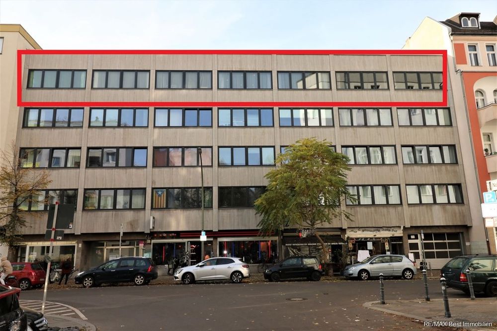 A bland, concrete, five-storey post-war building, with seven groups of four windows each on each of the upper floors, retail space on the ground floor. The top storey is outlined in red.