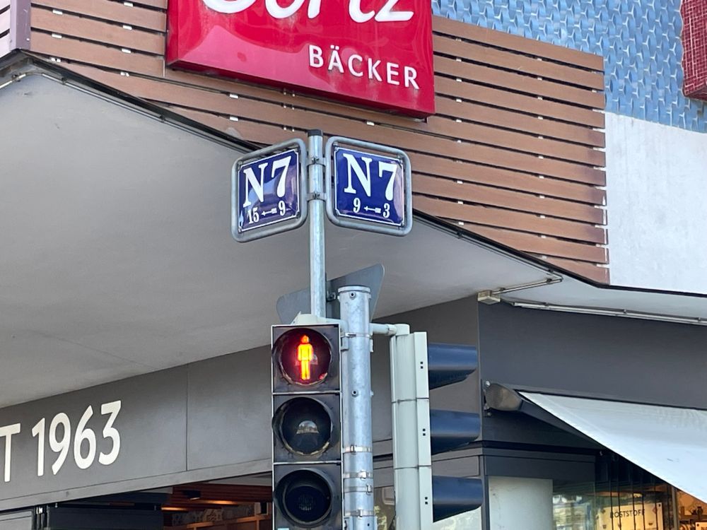 On a corner, two street signs in blue enamel with white text announcing this block in Mannheim as “N7”. Below it is a pedestrian traffic light on red, above it a sign saying “BÄCKER”.