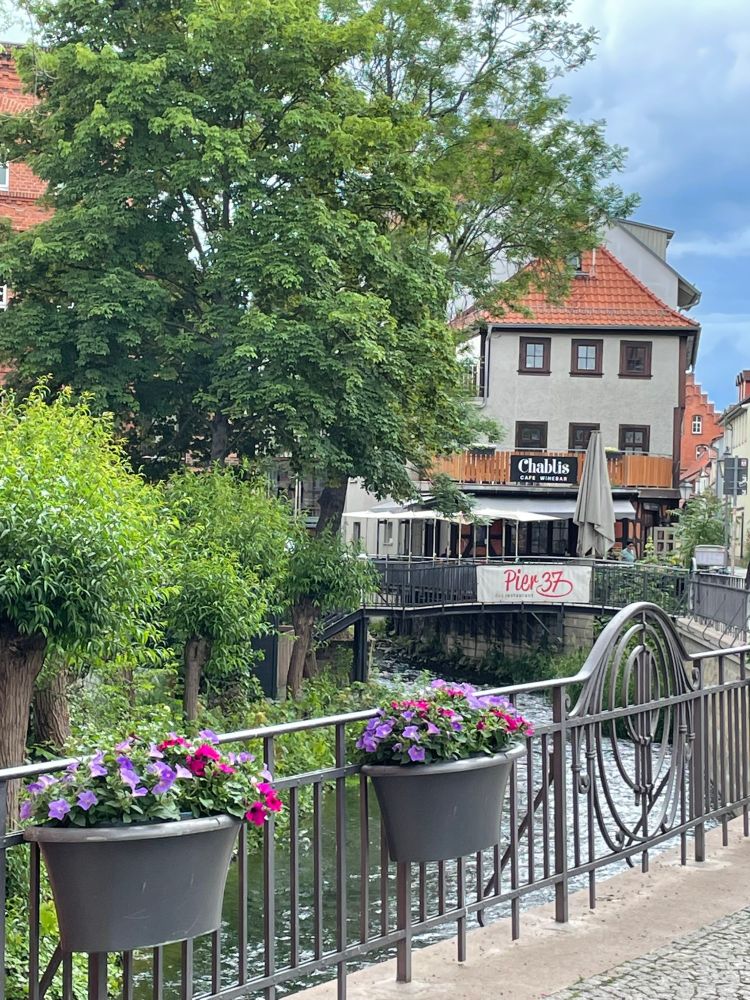 Narrow canal with railing from which hang potted flowers, beyond a restaurant overlooking the water; trees and greenery on the left side of the image with a brooding cloudy sky beyond.