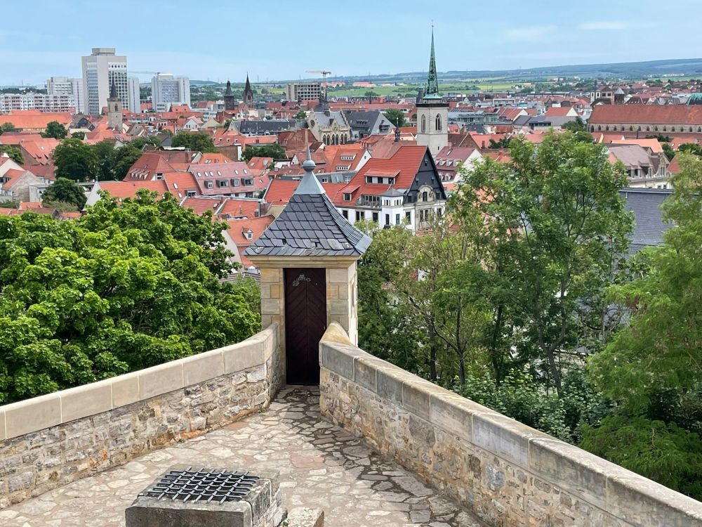 Looking down from a fortification with a terrace and a watchtower, with historic houses in the distance, modern apartment buildings further away, and green hills in the distance, under a largely blue sky.