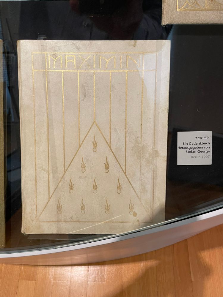 In a curved glass case, a book in a Jugendstill design with the title MAXIMIN in gold within a gold design with thin vertical lines, a triangle and flame-like motifs against an ivory-coloured background. To the right of the book, a label: "'Maximin'/Ein Gedenkbuch/Herausgegeben von/Stefan George/Berlin 1907"
