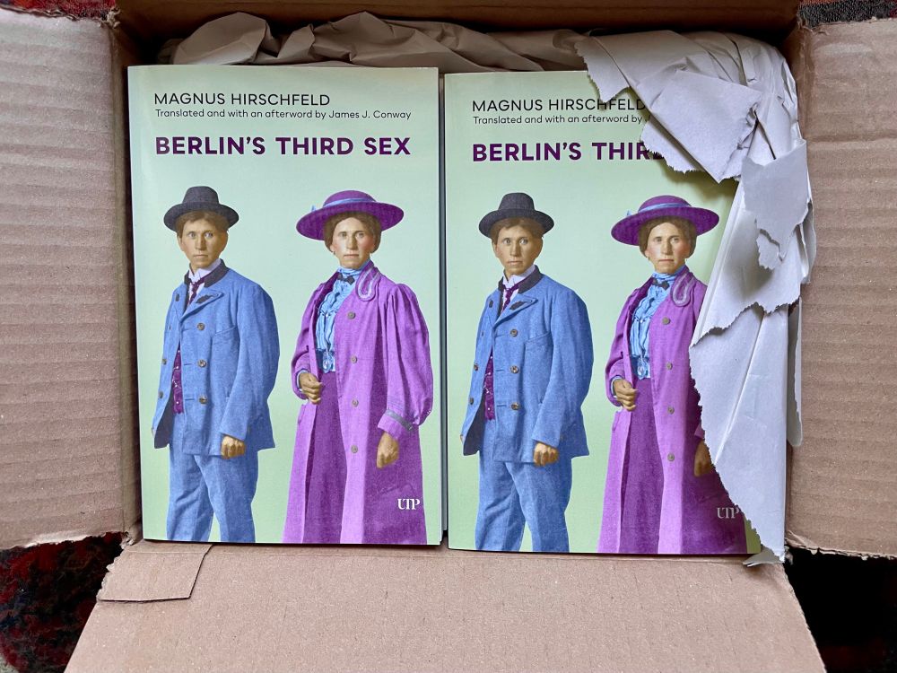 Cardboard box open to reveal two stacks of the same book. The cover of the book has a backdrop the colour of a green carnation, with two colourised photos showing the same individual in two different exposures, one in masculine-coded clothing from the early 20th century, and one in feminine-coded clothing. On the left the individual is wearing a suit coloured mid-blue with a white starched collar and purple tie and waistcoat and a black hat. On the right the individual is wearing a pink overcoat with wide sleeves over a light-blue blouse and a purple skirt, as well as a purple broad-brimmed hat with a blue ribbon. The text at the top reads MAGNUS HIRSCHFELD/Translated and with an afterword by James J. Conway/BERLIN’S THIRD SEX