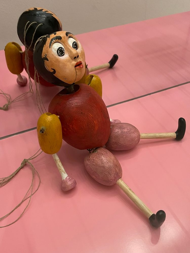 Stringed puppet figure with a dark red spherical torso, bulbous pink thighs and orange upper arms, stick forearms and lower legs, black hair with eyes agog, against a pick background