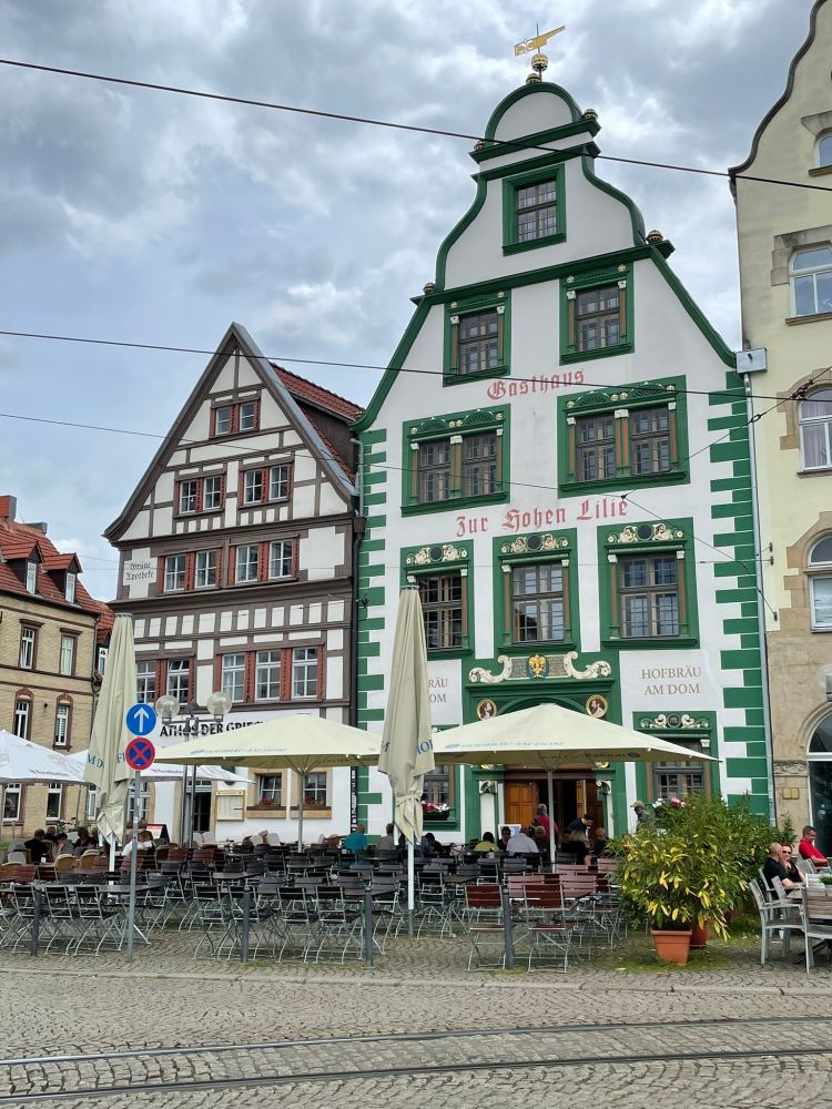 A public square with cobbles and tram tracks, with two historic buildings: on the left, a smaller half-timbered brown and cream five-storey building with a simple gabled roof, on the right a five-storey tavern in green and white with a bell gabled roof, behind them a cloudy sky.
