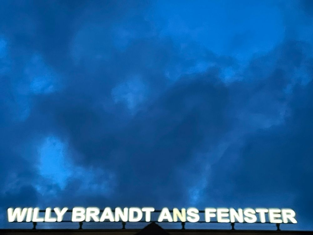 Brooding early evening sky, at the bottom of the image white neon letters spelling out WILLY BRANDT ANS FENSTER (Willy Brandt to the window).