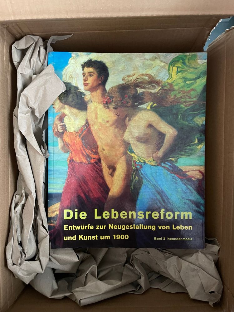 An open box showing a book with the title "Die Lebensreform/Entwürfe zur Neugestaltung von Leben und Kunst um 1900" in green against an oil painting showing a naked young man flanked by a bare-breasted young woman with long auburn hair and green cloth blown by the wind and a blue skirt, and a dark-haired woman with an orange top and red skirt with her head turned away. They look to be striding into a better future. 