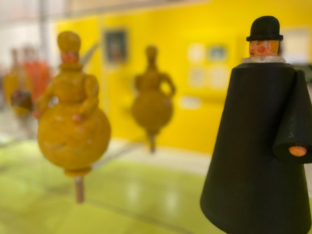 Toy figures in a cabinet; a conical black-clad figure in the foreground, a spherical figure out of focus further away against a green and yellow background