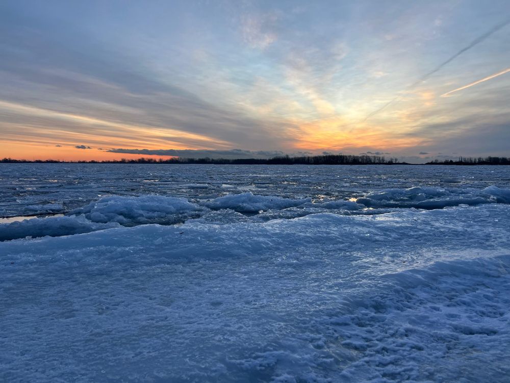 Frozen lake solid in the foreground, ice chunks middle ground orange sun coming up under high clouds.