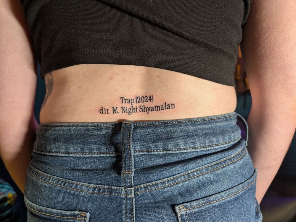 A picture of Farky from behind. She's wearing a black crop top and jean shorts. In the space between, on her lower back, is a tattoo of the words "Trap (2024) dir. M. Night Shyamalan" in a serif font