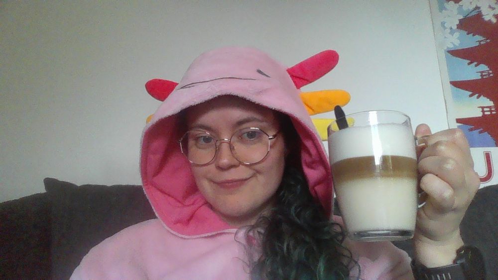 Une photo portrait de Globulle, un macchiato à la main, habillée d'un superbe kigurumi axolotl. Elle porte ses lunettes et fait un petit sourire, mais la fatigue se lit dans ses yeux