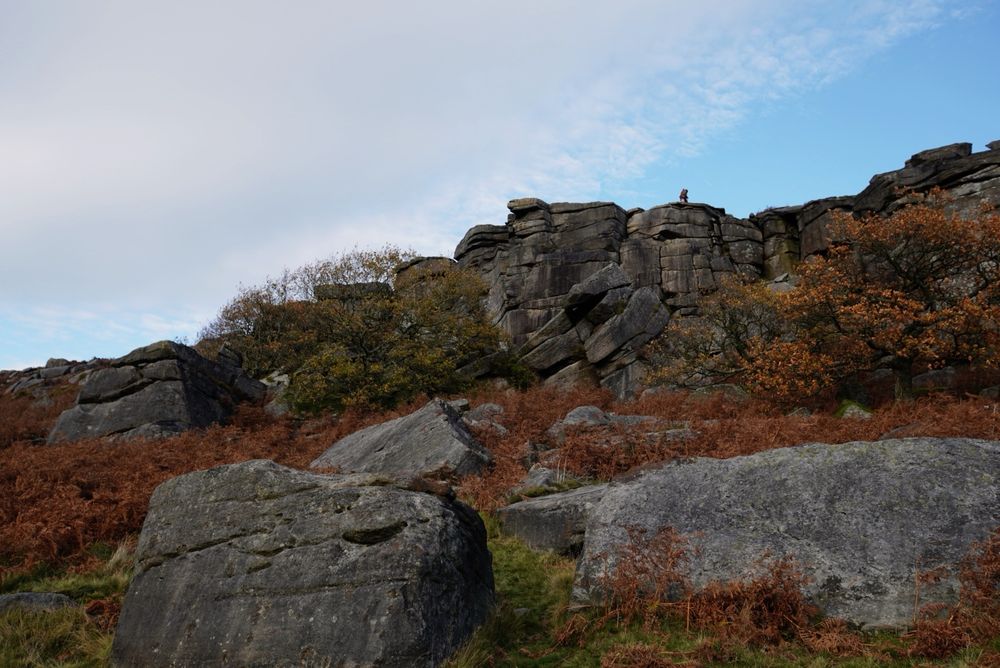 A climber is framed against the sky on top of a gritstone edge. Trees with yellow and orange foliage crowd at the bottom