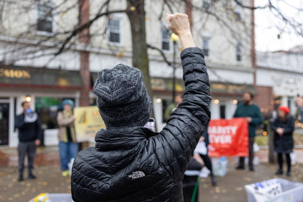 Photo of a woman taken from behind. She’s wearing a black jacket and hat and her fist is up in the air.