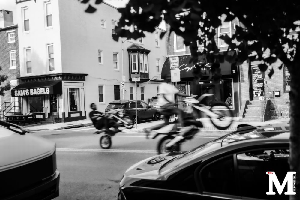 Two dirtbikes ride past “Sam’s Bagels”. M “the shooter” is in white in the bottom right hand corner. 