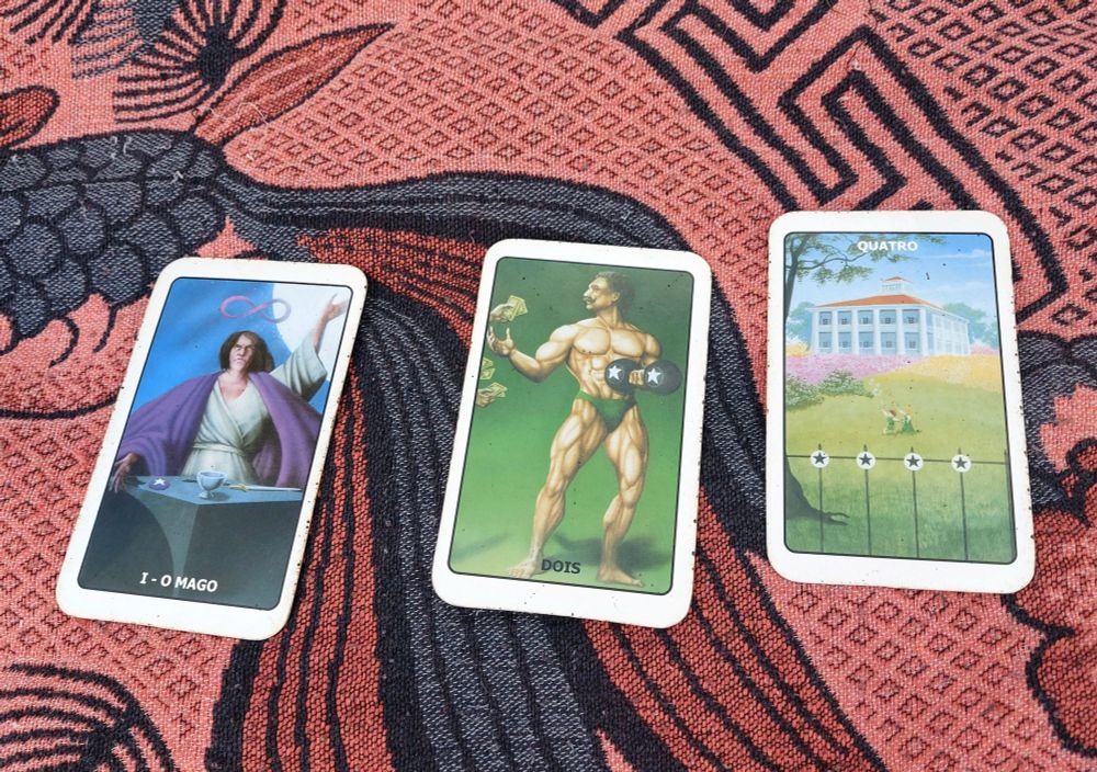 Três cartas do Tarô das Bruxas em cima de um forro em tons de rosa e preto. As cartas são "O Mago", "Dois de Ouros" e "4 de Ouros".