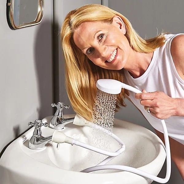 a smiling white blonde woman using a shower attachment that fits on to hot & cold taps so she can apparently have a shower in the washbasin 