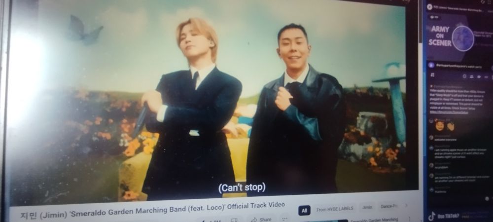 A streaming picture from BTS Scener. In the picture, Jimin wearing a Black suit with Loco and in a fantasy-like background. This is from the music video of his song Smeraldo Garden Marching Band.