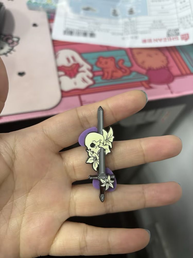 A hand holding a pin depicting a sword impaling a skull through its mouth. There are two Lilly flowers around the skull and another on the handle. 