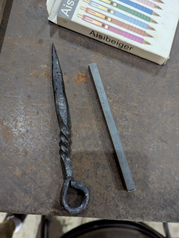 A rectangular stick of steel next to a kunai inspired letter opener which has been made from a similar piece of steel
