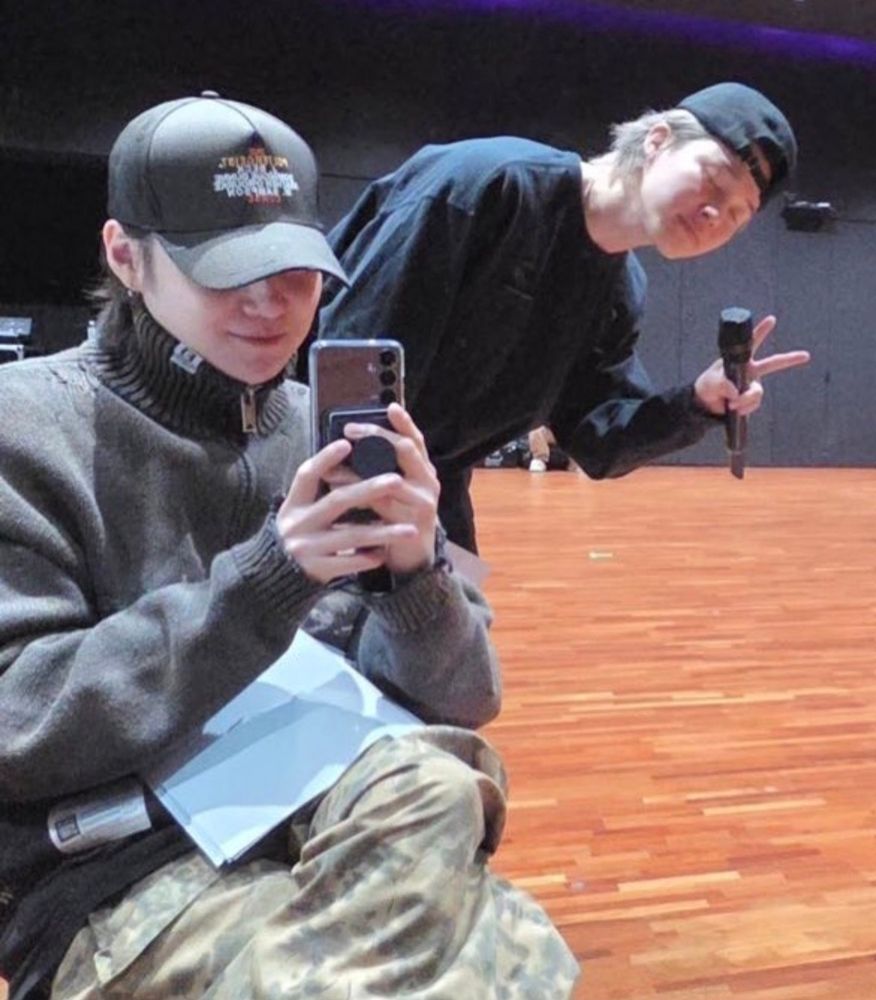 Yoongi taking a mirror selca in the dance room, with Jimin poking out behind him with a peace sign. Yoongi's cap obscures his face apart from his cute, tiny smile.