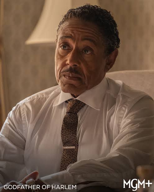 Foto em plano médio, em tom de sépia ,de Giancarlo Esposito sentado, caracterizado como o pastor Adam Clayton Powell Jr para a série Godfather of Harlem, vestindo camisa branca e gravata escura.