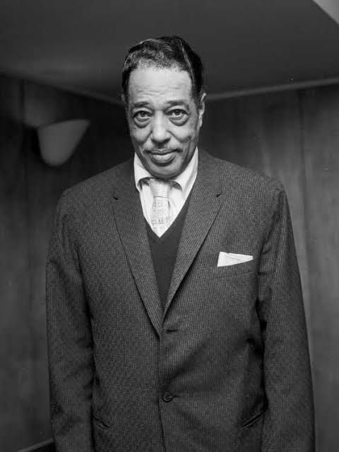 Foto em preto e branco em meio corpo de Duke Ellington, de pé, vestindo terno e gravata encarando a câmera.