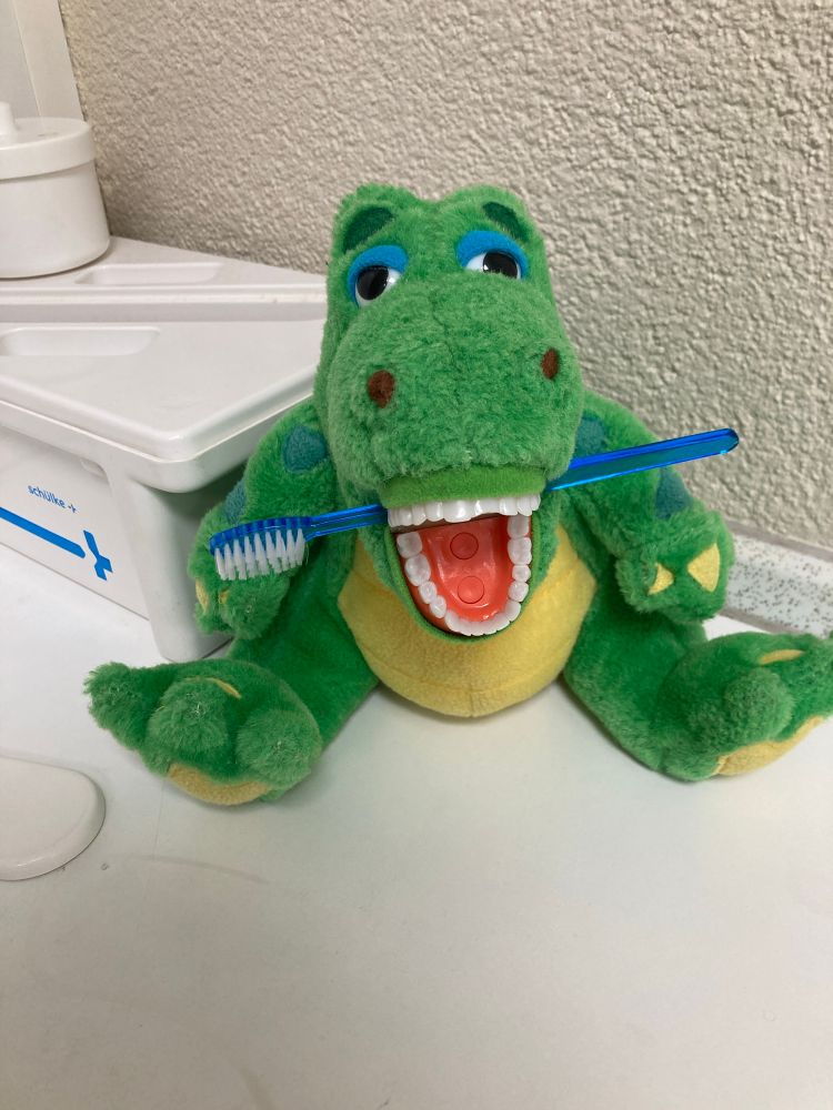 Photo of a crocodile plushie with a plastic set of human teeth as a mooth. A toothbrush is stuck in between the teeth. 
