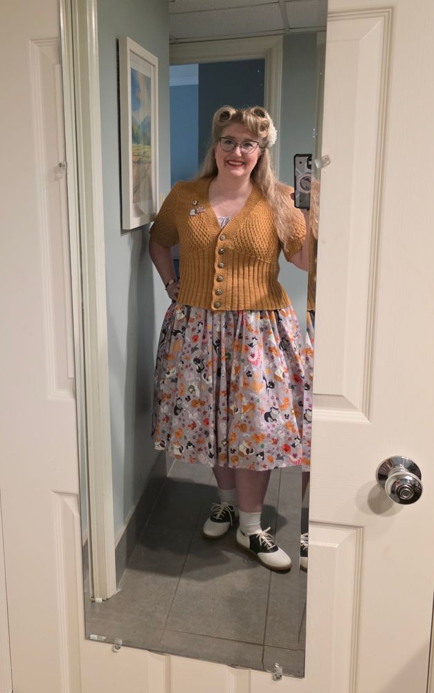woman in disney cats dress with a gold colored short sleeve cardigan. Her hair is in victory rolls and she is wearing saddle shoes.