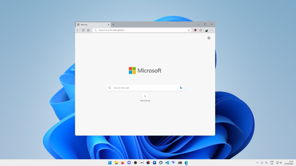 A bare-looking Microsoft Edge window atop a Windows 11 desktop, which features the typical default Bloom wallpaper in light theme. Pinned to the taskbar are File Explorer, Firefox, Discord, OBS, ChroMapper, BSManager, the PlayStation VR2 App, SteamVR, Visual Studio Code, ProtonVPN, and the Windows Terminal application; Edge sits to the right of the latter on the taskbar, and Discord's system tray icon sits to the right of that, beside the arrow to reveal more system tray icons.

The taskbar displays an indicator reading 'ENG', then, on a new line, 'UK'.

The taskbar indicates that this computer is connected to a network via a wired connection, and that the time is 15:31, on the date 06/08/2025, meaning the 6th of August 2025.

Edge lacks much on the top bar, displaying only a back button, a reload button, and a home button, alongside a search bar that reads 'Search or enter web address', with an empty favourite button within it to the very right. Next to this is the uBlock Origin icon, symbolising activity with its red shade, and beside this is the extensions button for opening the menu through which extensions not 'pinned' to the top bar may be accessed. Afterwards, there is a user profile icon, taking the form of the  profile picture of the currently signed in user: a shot from the side of the helmet of a robot from Hyper Dash. Then, the three dots menu is present to the  very right edge of the top bar, allowing for more options to be accessed. The New tab page displays a 'Microsoft' logo as is typical of the Edge New tab page while the 'Background' option is disabled. No content is enabled on the New tab page, only this logo, a Bing search bar and an icon to 'Add [a] shortcut' is visible, with a settings cog to the top right corner.